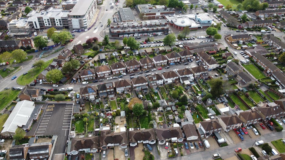 An aerial view of a suburban residential area showing a row of terraced houses with small, well-maintained gardens featuring lush green lawns, trimmed hedges, and a variety of plants and trees. The ga
